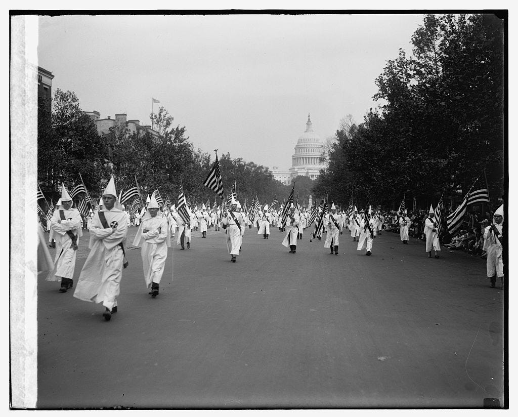 Black and white image of Ku Klux Klan members in white robes holding US flags marching with the US Capitol visible in the background.