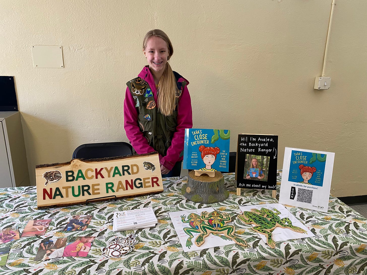 photo of a young girl with long blond hair and a purple shirt wearing an olive green vest decorated with wildlife related badges. She stands in a room behind a table with a sign that says Backyard Nature Ranger, an image of her book cover (Tara's Close Enouncter," a picture book) and stickers showing reptiles. The table is covered with a tablecloth that has green leaves and other nature designs. She is in a function room with pale yellow cinderblock walls. 