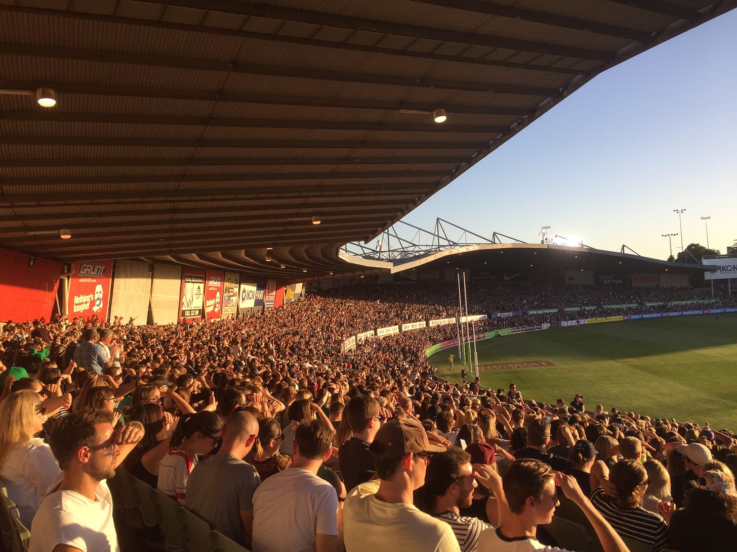 A photograph of a fully packed crowd at Princes Park stadium. They are all bathed in the golden glow of the setting sun. Many people are holding the hands up to block the sun. You can see the edge of the green field with the goalposts.