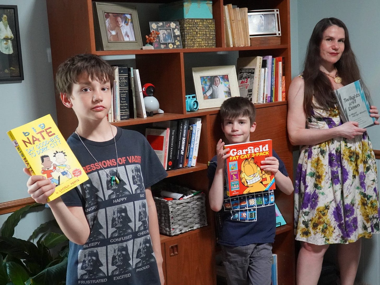 In front of bookshelf full of books and photos, stands a boy (William Hidalgo-Bowler) holding a Big Nate Say Good-Bye to Dork City book, another boy (Santiago Hidalgo-Bowler) holding a Garfield Fat Cat 3-Pack book, and a woman (Alexandra Hidalgo) holding the book Twilights and Dawn by Miguel Hidalgo. They look at the camera. It is daytime. In front of bookshelf full of books and photos, stands a boy (William Hidalgo-Bowler) holding a Big Nate Say Good-Bye to Dork City book, another boy (Santiago Hidalgo-Bowler) holding a Garfield Fat Cat 3-Pack book, and a woman (Alexandra Hidalgo) holding the book Twilights and Dawn by Miguel Hidalgo. They look at the camera. It is daytime.