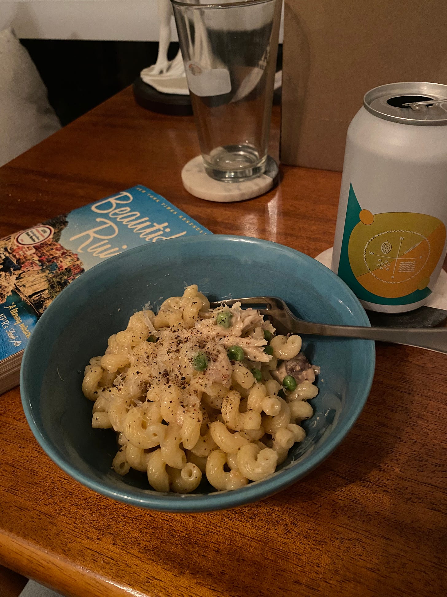 A blue bowl of short noodles in a creamy sauce with pieces of shredded chicken, green peas, and lots of black pepper. Behind it is a paperback novel, "Beautiful Ruins", and to its right on a coaster is a can of Four Winds beer.