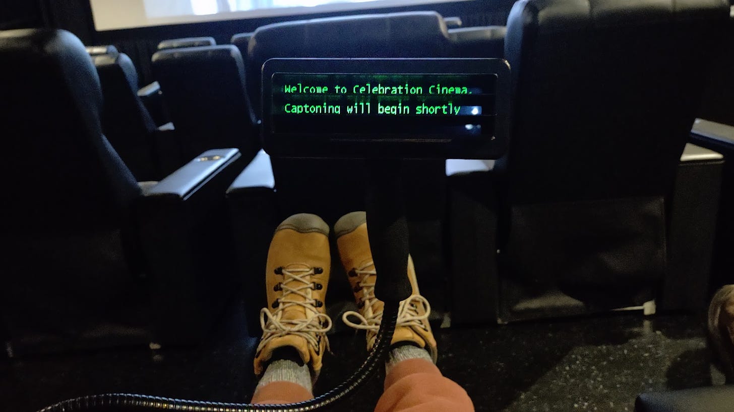 A closed captions device that shows the text "Welcome to Celebration Cinema. Captioning will begin shortly. The background shows cinema theater seats and screen.