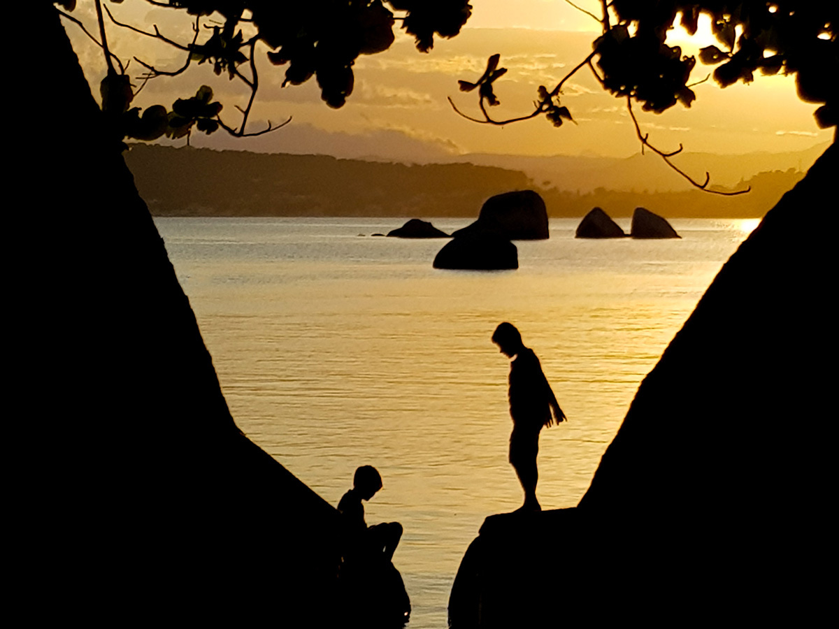 Foto colorida ao pôr do sol com o céu e o mar dourados, com duas crianças sobre pedras no primeiro plano e diversas outras pedras no mar ao fundo.