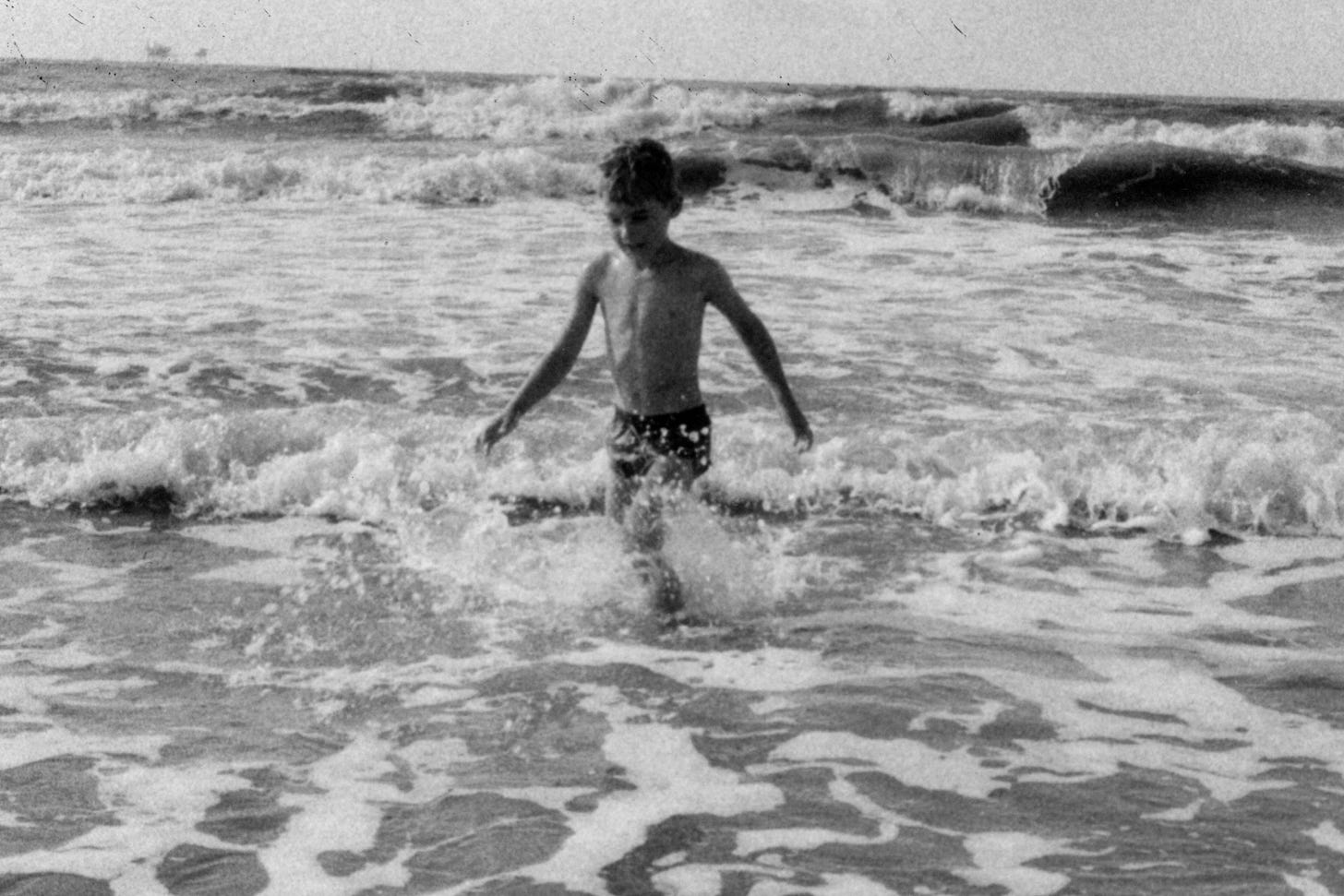 Black and white photo of a boy young boy standing in the rolling waves smiling in delight