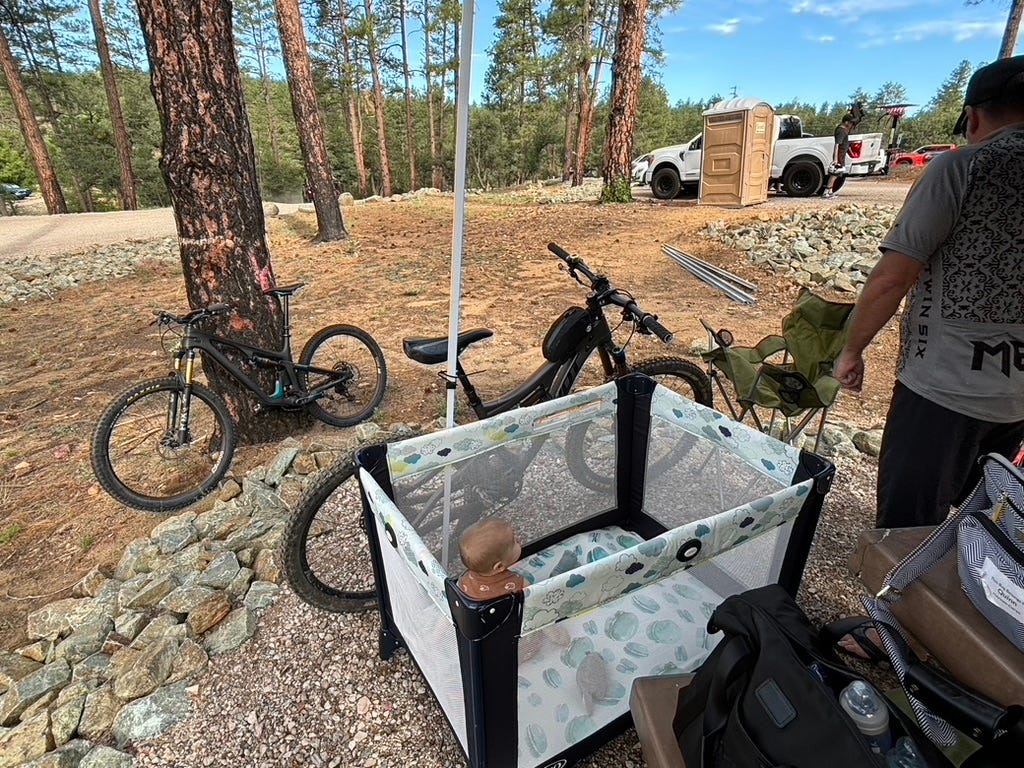 A camping scene featuring two bicycles parked next to a playpen with a small child inside. In the background, there are trees, a portable toilet, and people setting up equipment. The ground is rocky and gravelly, typical of an outdoor recreational area.