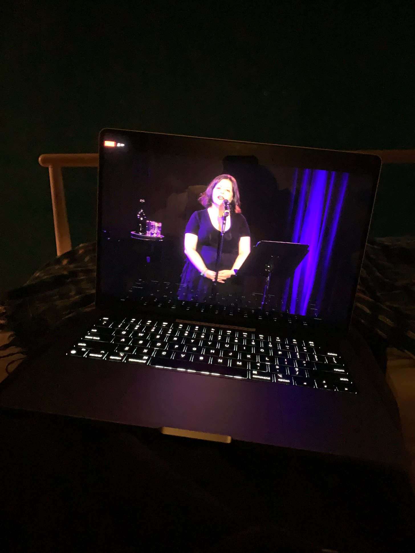A white woman dressed in a black dress stands on a stage behind a microphone, singing. This image is on a computer sitting on the lap of a person in a dark room.