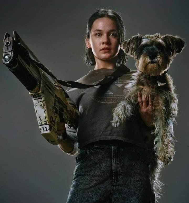 Image of lead female character holding a huge gun in one hand and a cute salt and pepper schnauzer dog in the other.