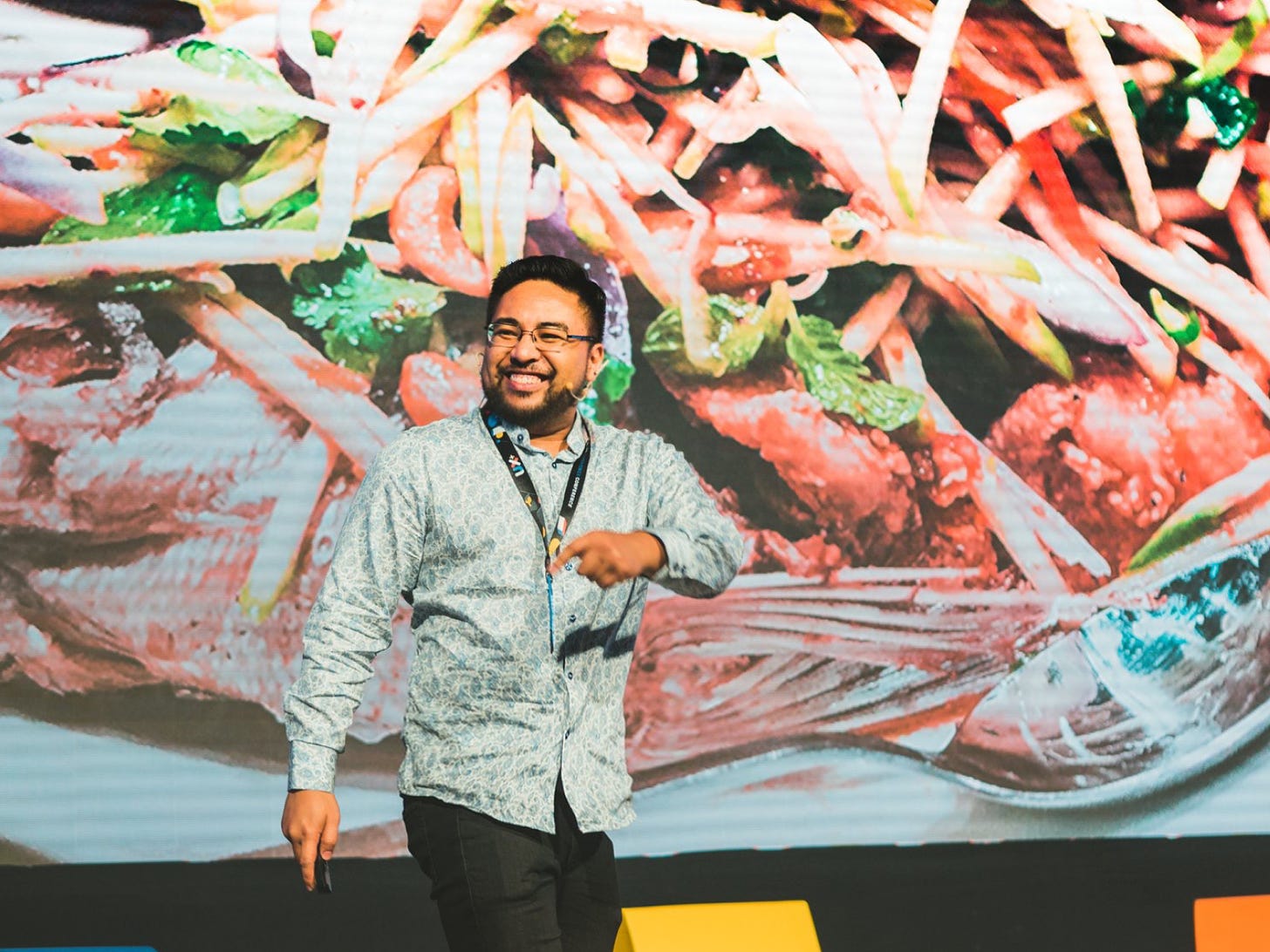 Jay Demetillo smiling and pointing on stage at the UX+ conference in front of a giant photo of food.