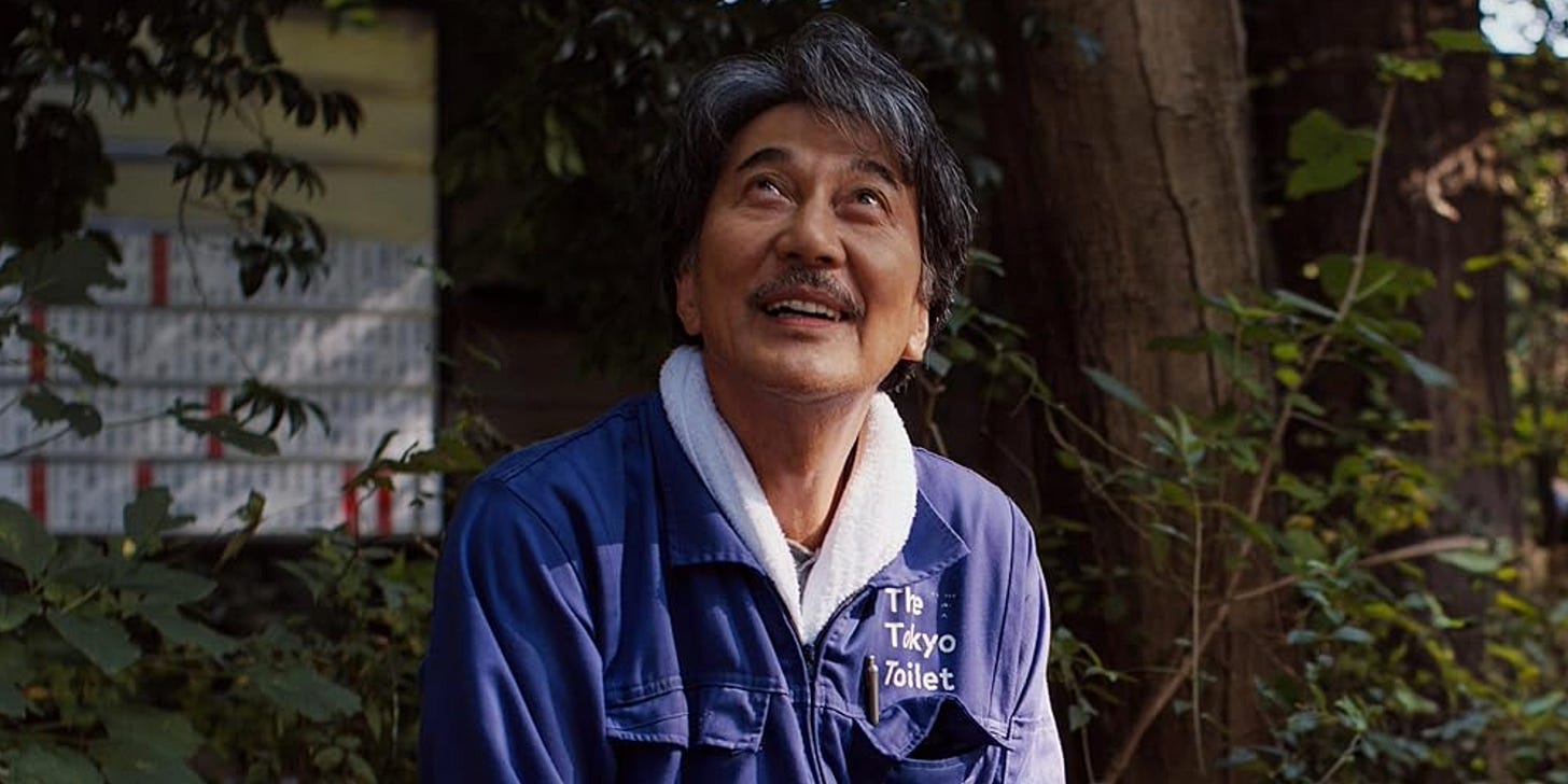 Hirayama, a middle-aged man, sits in his blue uniform underneath some trees. He looks up and smiles at the sky.