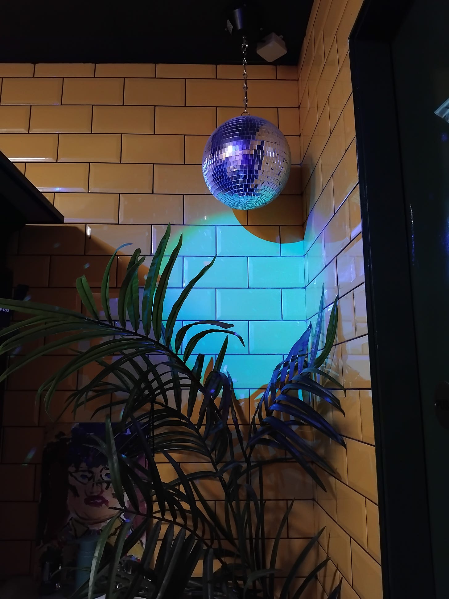 In the right corner of a room, a tiny disco ball shimmers with reflective lighting. The wall has yellow tiles, and a palm type plant sits in a pot below it. 