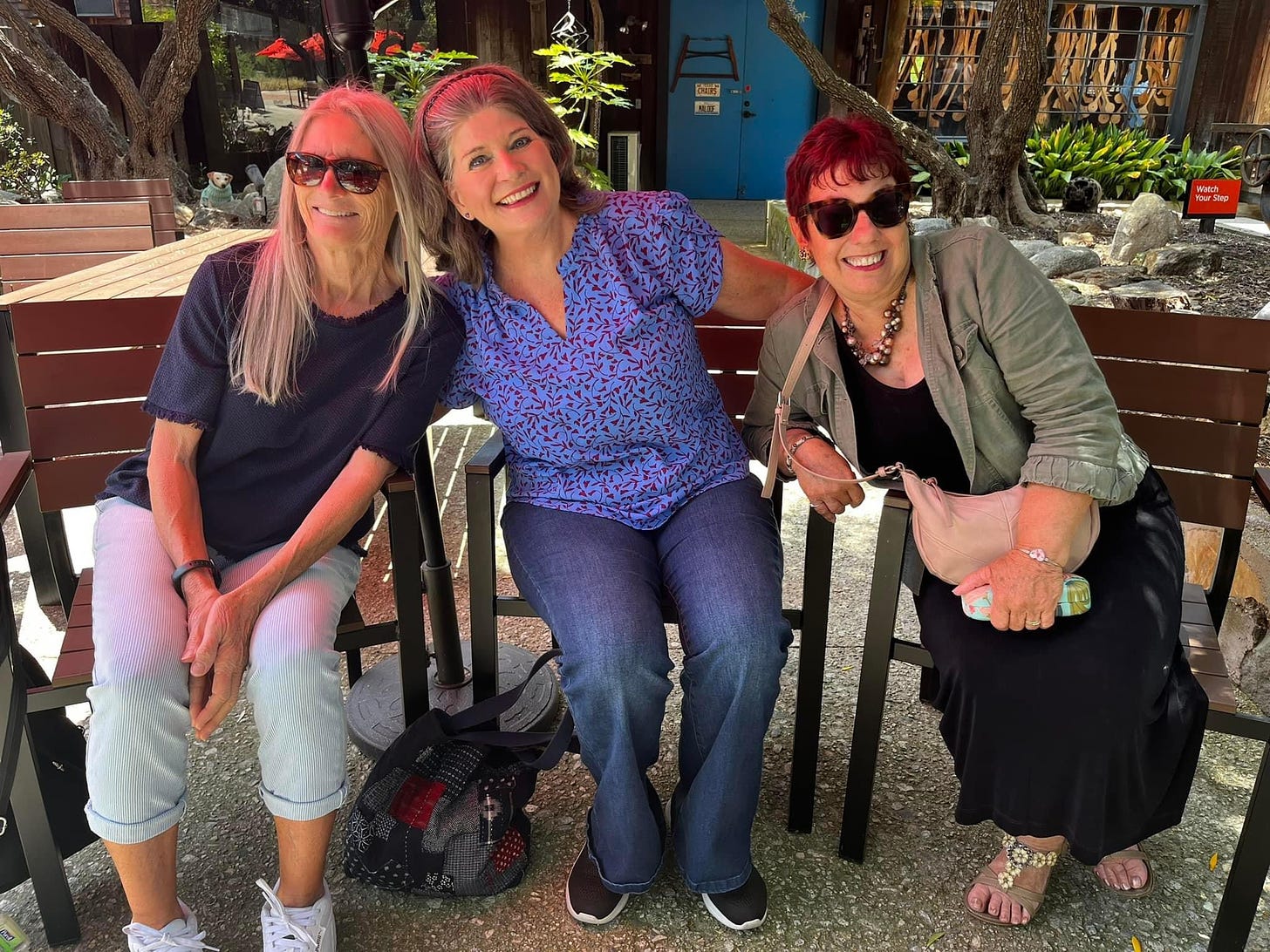 Three women sitting on wooden chairs outdoors and smiling for the camera.