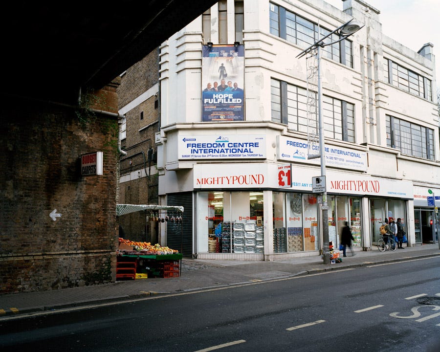 Photo by Chloe Dewe Mathews from a series “Sunday Service” showing an empty Rye Lane, a street in South London known for its large African and Caribbean communities. The view is from under a railway arch with Might Pound discount store at its centre and a sign to an African church “Freedom Centre International” signposted to the side.
