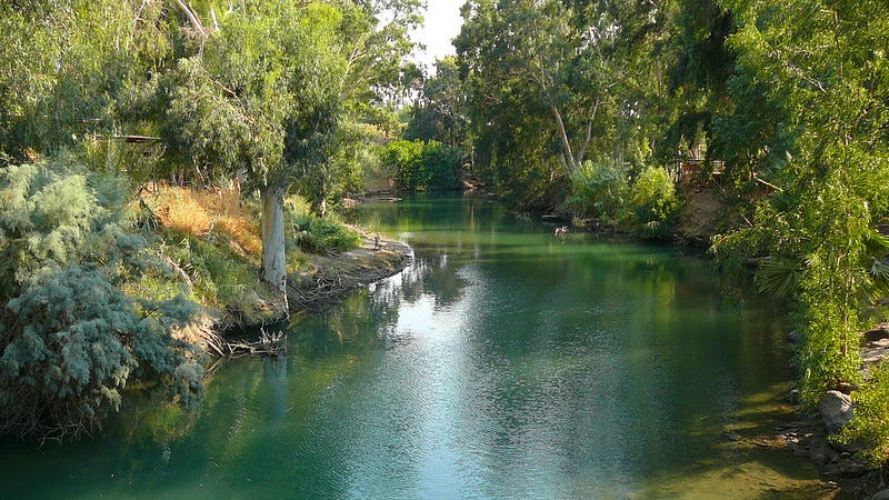 A bend in the Jordan River with green, gently rippling water surrounded by much vegetation.