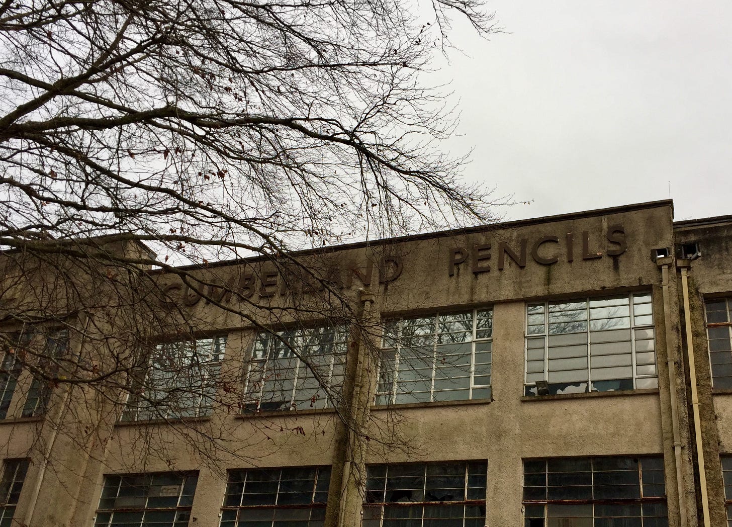 Photograph of the front of a factory, block letters on the front read Cumberland Pencils. The building is disused with a few broken windows. A bare winter tree is in the foreground