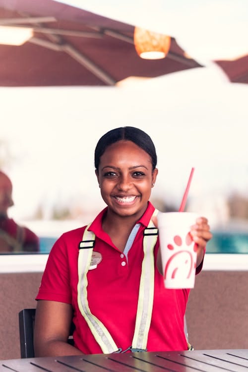 Free Cheerful employee in uniform holding a branded cup with a smile at an outdoor cafe table. Stock Photo