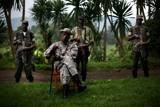 Rebel leader Laurent Nkunda poses outside for a portrait sitting on a chair and surrounded by 3 armed guards.