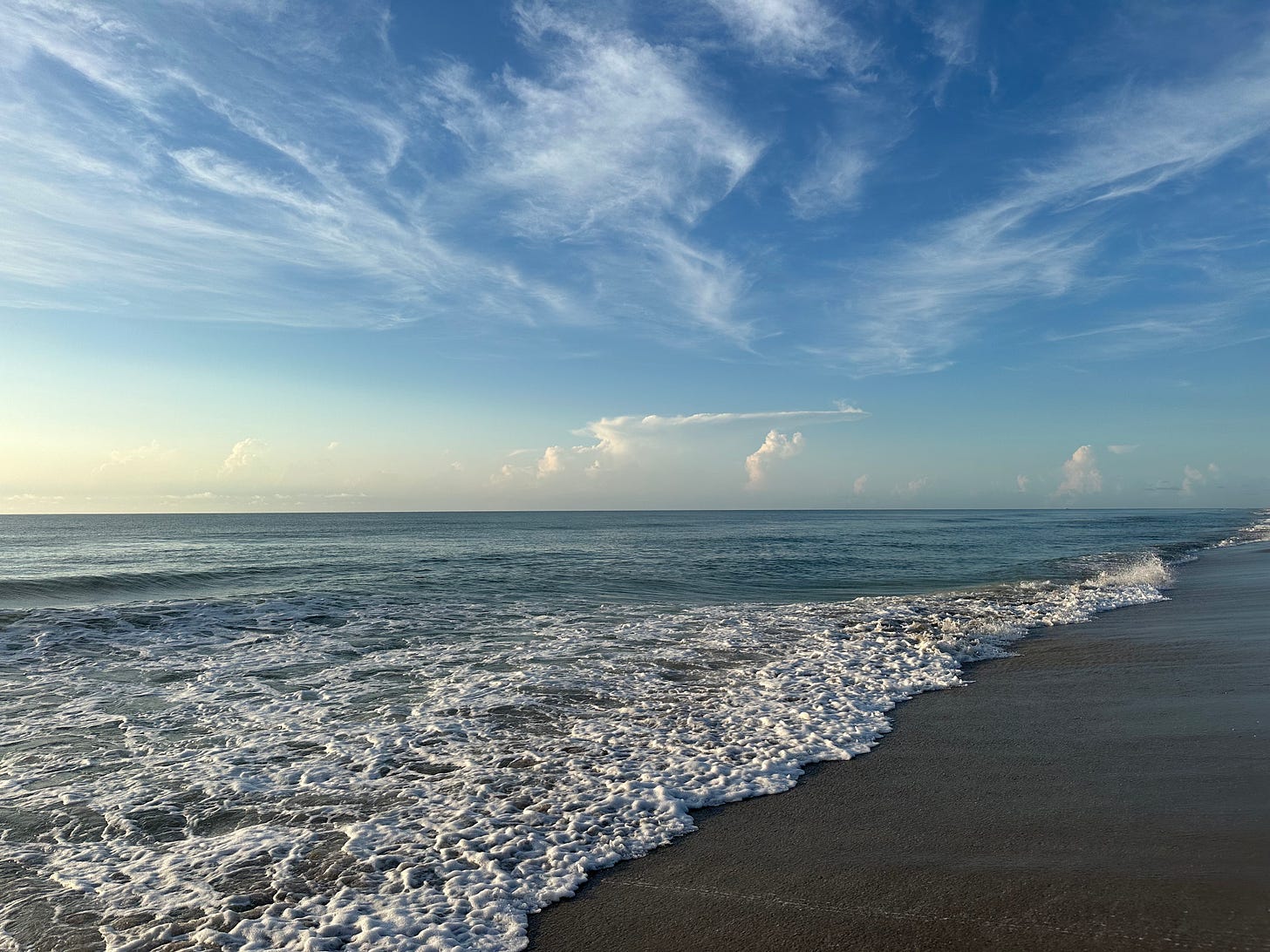 A photo of the ocean shoreline with a bright blue sky and setting sun above