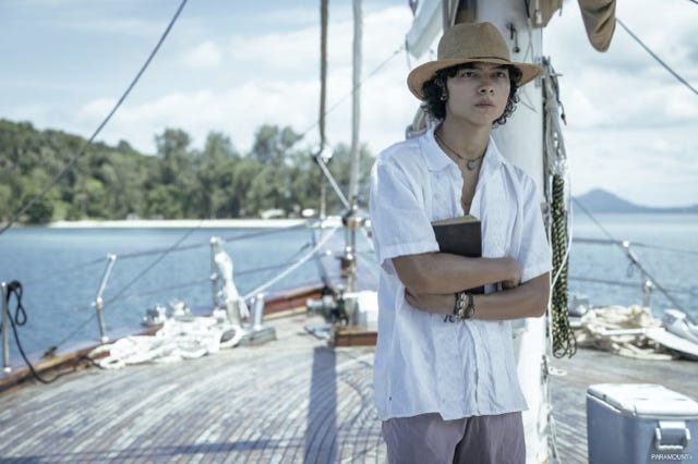Narayan Hecter in character in a scene from No Escape, he stands in a white shirt and hat on a boat