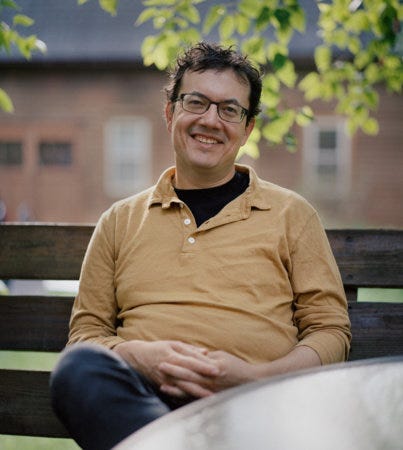 A photograph of author Andrew Leland. Sitting on a park bench, smiling with a mustard-yellow shirt. He's a middle-aged white man with his fingers templed.