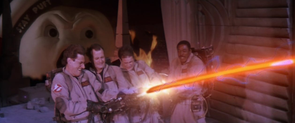 Still from Ghostbusters, where Egon, Peter, Ray, and Winston cross the streams of their proton packs with the Stay Put Marshmallow Man coming up behind them.