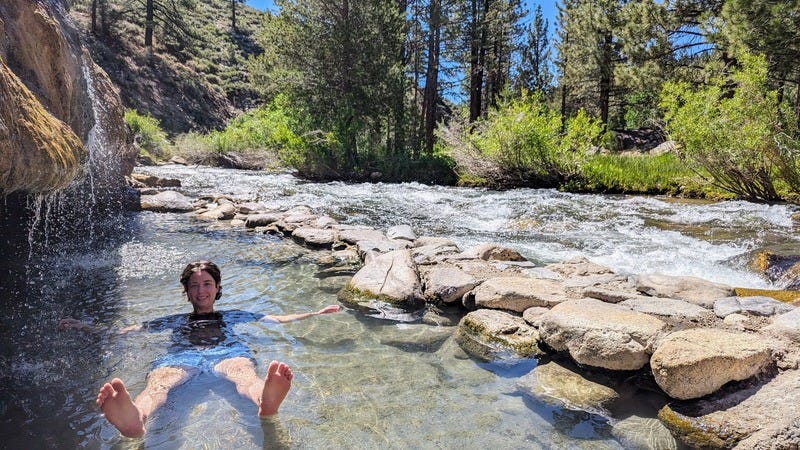 Rey, a white nonbinary person wearing a t-shirt and swim trunks, floating in a hot spring pool with a small waterfall on the left and a rushing stream on the right