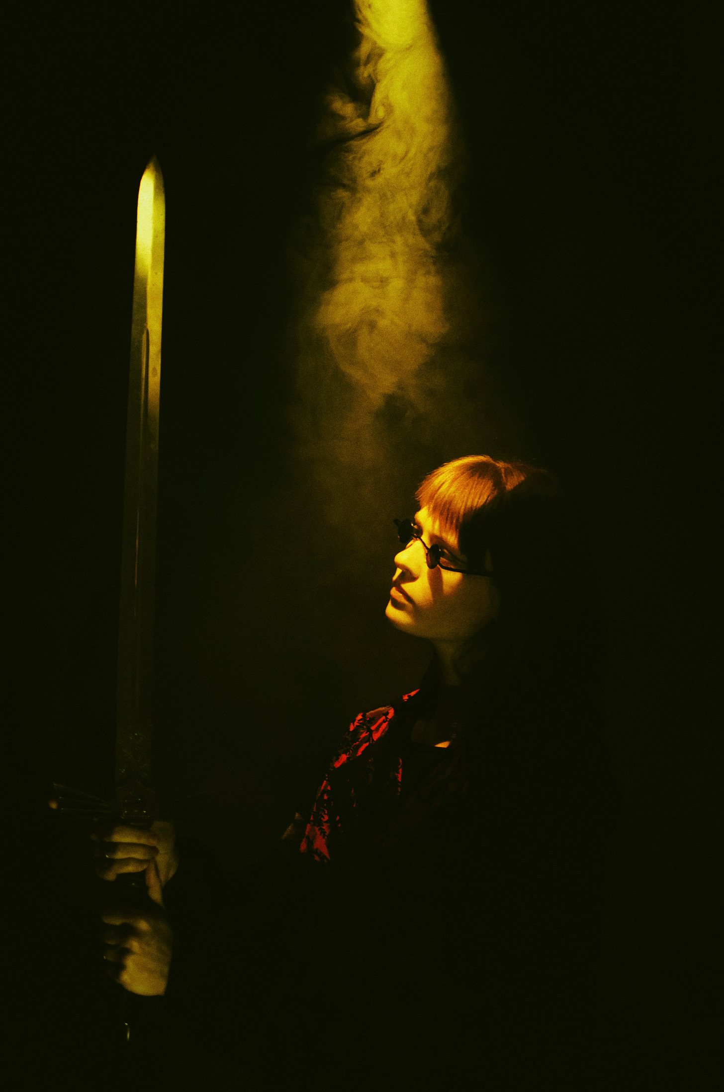 A red-haired person in small dark sunglasses with a bright red scarf at their neck holds a long sword upright in near darkness. Smoke swirls behind them.