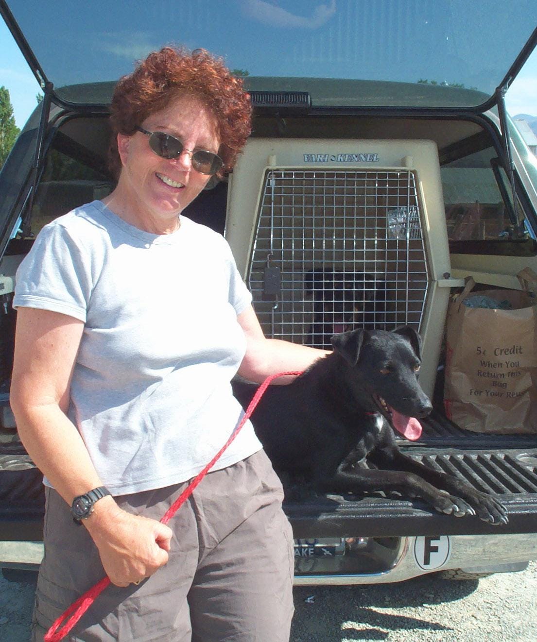 Photo of a smiling red-headed woman holding a smiling black lab on a loose leash. They are at the rear of a hatchback vehicle with a large animal carrier.