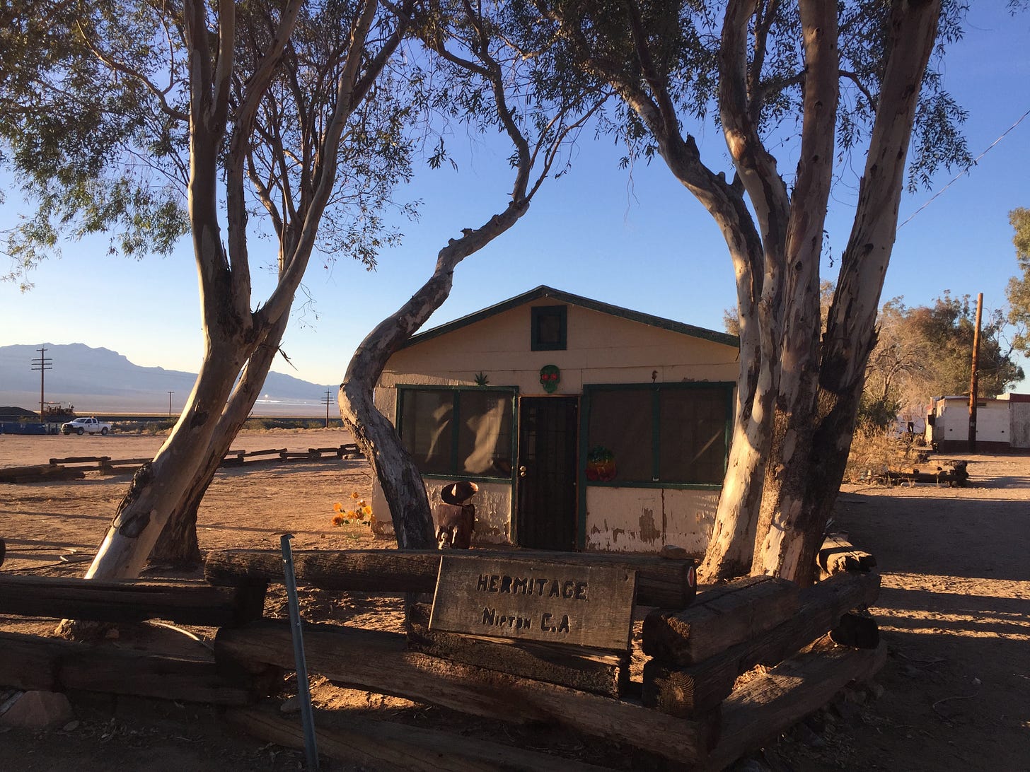 A ramshackle white house with green trim beneath eucalyptus trees in the Mojave Desert.