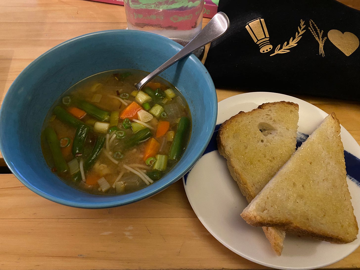 The miso vegetable soup described above, in a blue bowl with a spoon in it. Next to it is a slice of buttered toast, cut in half on a white plate with a blue swipe design. Behind it are a glass of water and a small black bag with a salt shaker, two herb sprigs, and a gold heart on it.