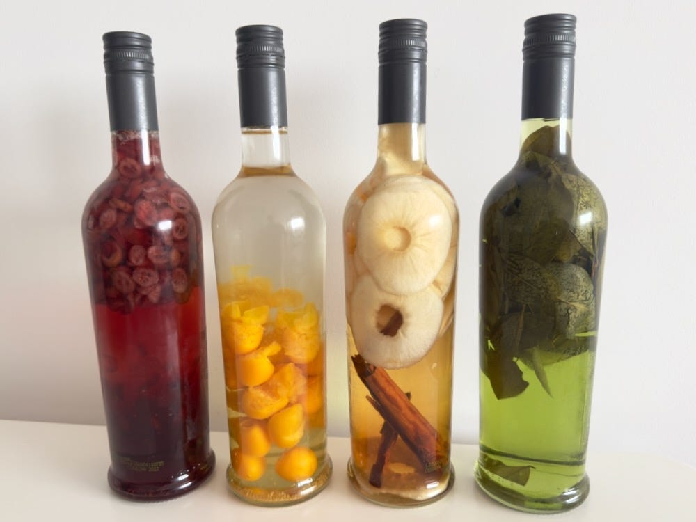 A row of four colorful bottles of macerados: cranberry (deep red), aguaymanto (orange yellow), apple cinnamon (light yellow), coca leaf (green)