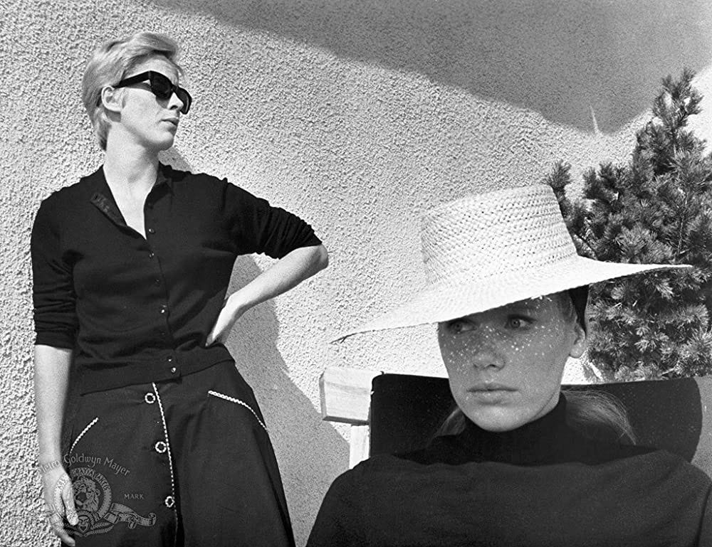 A still from the black-and-white film Persona. In the background, Bibi Andersson, a woman with short blond hair wearing sunglasses and a dark colored sweater, stands with one hand on her hip looking to her left, where Liv Ullman, a woman with long hair wearing a wide-brimmed hat, sits in the foreground.