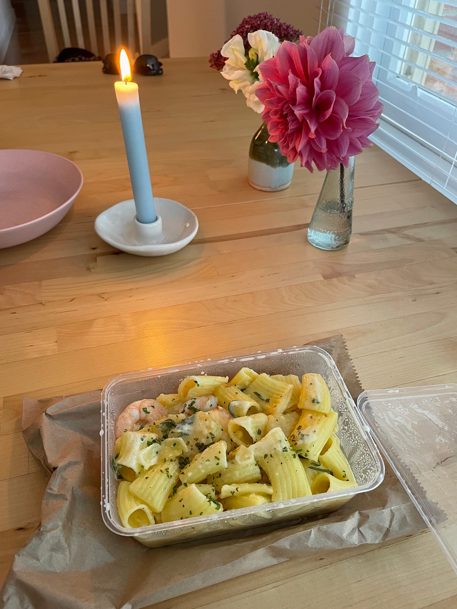 Candle at dinner time with dahlias and a takeaway container of pasta.