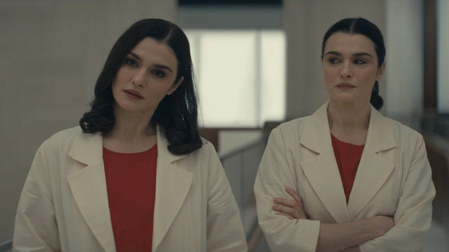 Two white women with dark hair stand next to each other. They are identical twins and are both wearing red shirts with white lab coats. The one on the left has her dark shoulder-length hair down and she looks forward with her head tilted and an inquisitive expression. The twin on the right has her dark hair parted in the middle and pulled back. Her arms are crossed and she looks seriously at her sister.