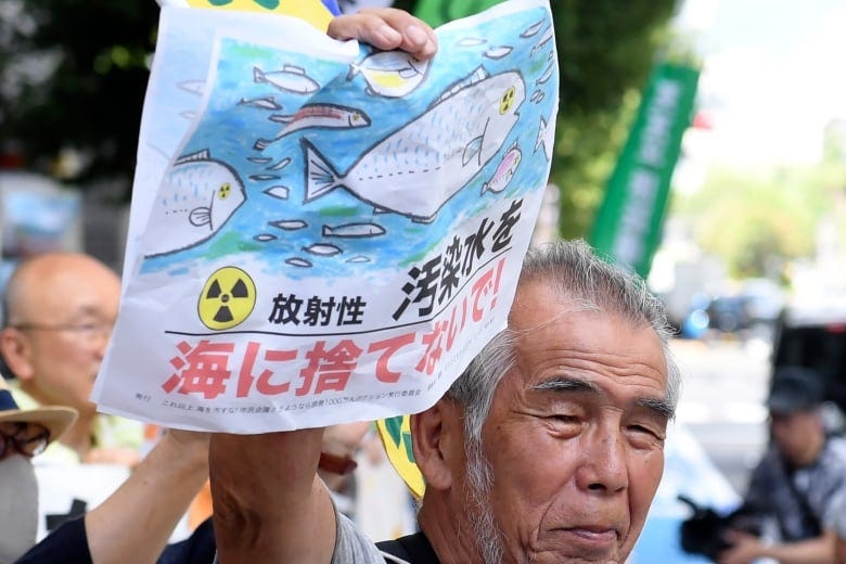 An older man holds a sign with cartoon fish, blue water and a radioactive symbol. The sign says, 'Do not discharge the wastewater into the sea' in Japanese.