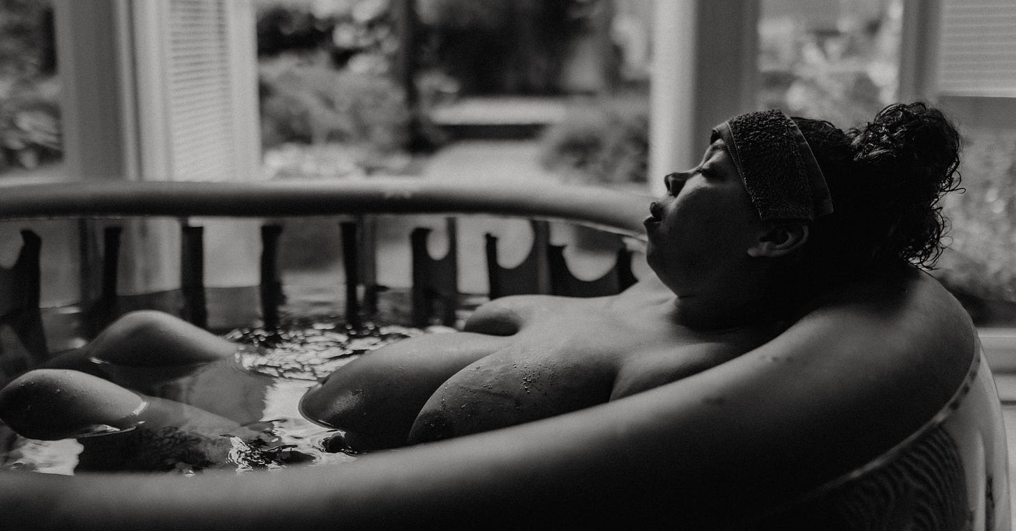 A pregnant person in labor, partially submerged in a birthing pool, leaning back with their eyes closed and a headband on. Soft natural light filters through a window in the background.