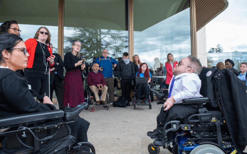 About 15 disabled people, several in wheelchairs, sit and stand in a rough circle while talking outside a glass-fronted building