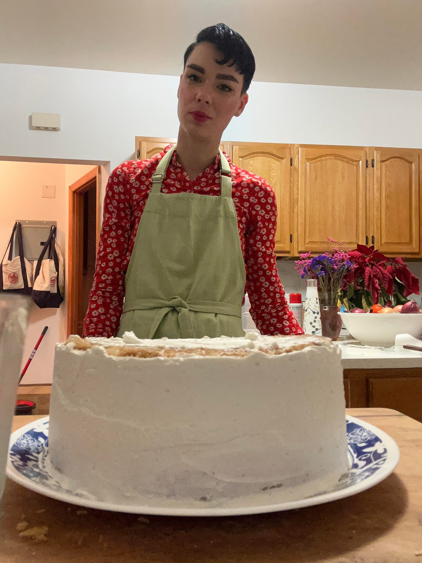White person with short black hair wears a red shirt and a green apron, they stand behind a half-frosted white cake in a large kitchen