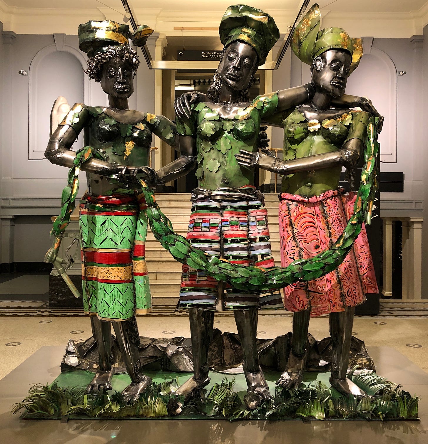 A sculpture of three metallic female figures stand in a row, on a pedestal simulating a grassy floor, in a museum gallery. The outer figures support the central figure. All three wear skirts and headdresses.