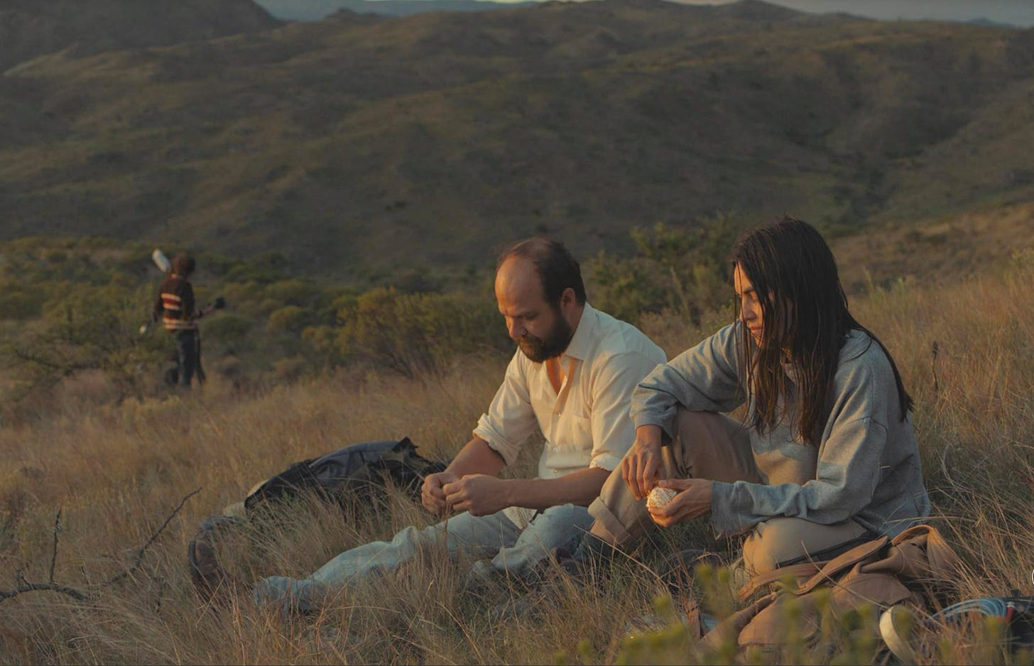 A man and a woman sit on a grassy ridge in late-afternoon sunlight. Beneath and beyond them are grassy hillsides and ridges filling the frame.