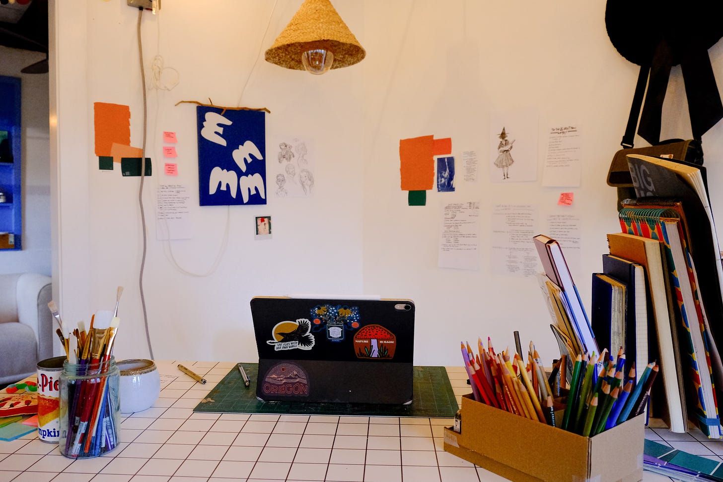 Widest of Gracie's workspace shows a rainbow of colored pencils, an iPad, art pinned to the wall, and a stack of notebooks and sketchbooks.