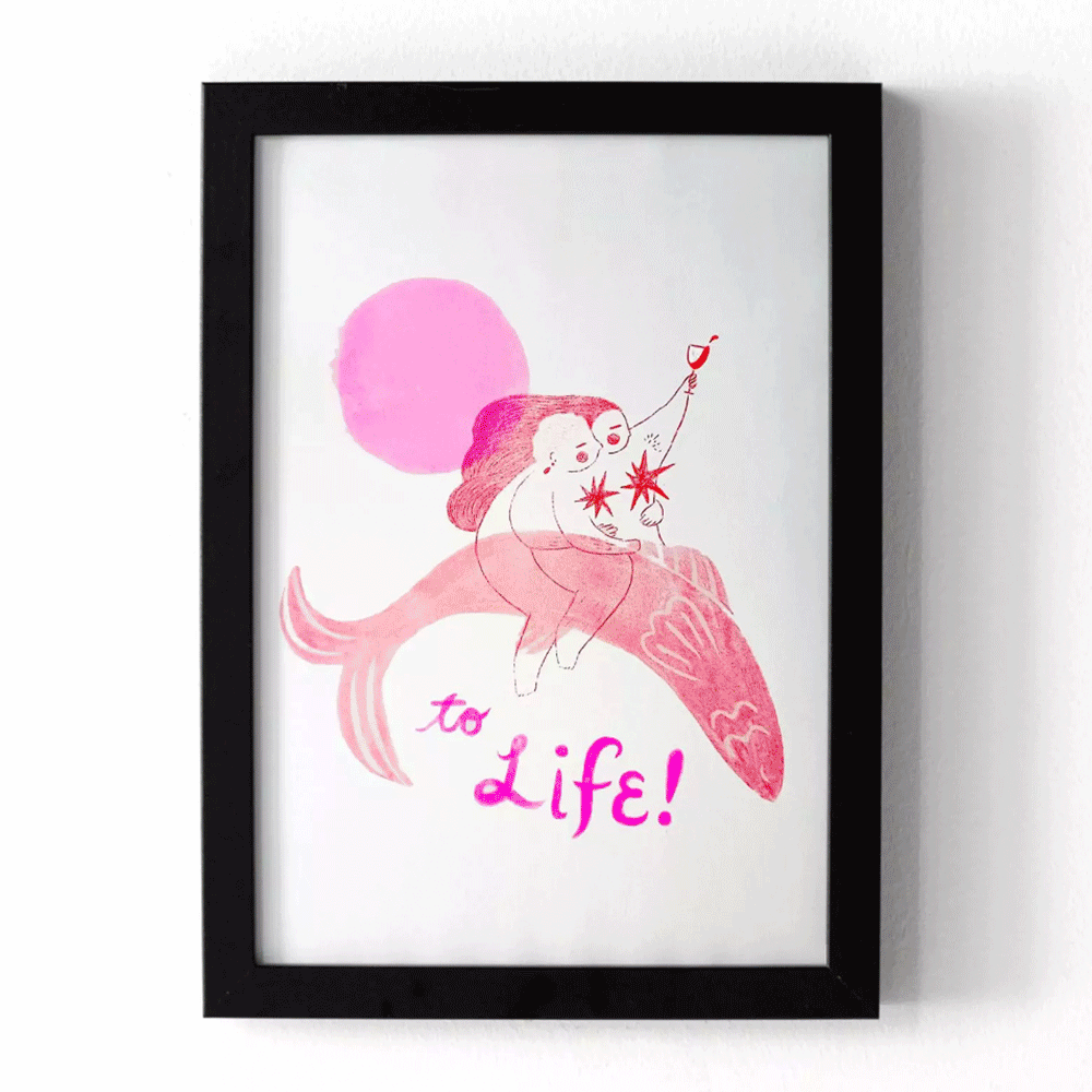 GIF featuring a pink poster of two people riding a fish celebrating life. The artist is also wearing pink and is signing their posters and posing next to a framed version of their print.