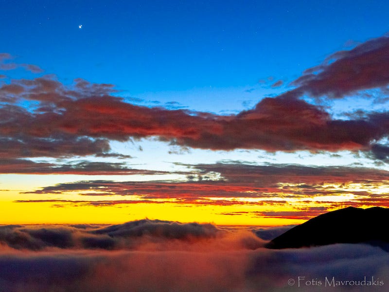 Wispy white in front of dark mountains, yellow and blue sky with red clouds and 2 bright dots in upper left corner.