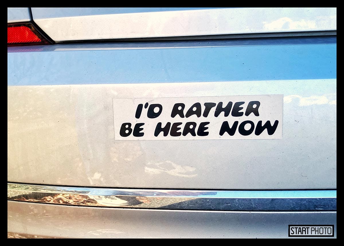 Color photograph of a bumpersticker with the text, "I'd rather be here now".