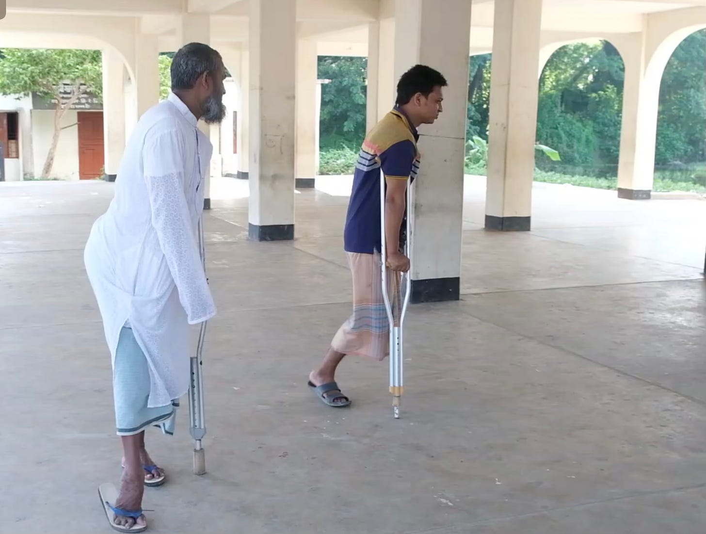 Two men stand in an open air pavilion. One balances himself with a crutch, another man with one leg studies himself with a crutch.