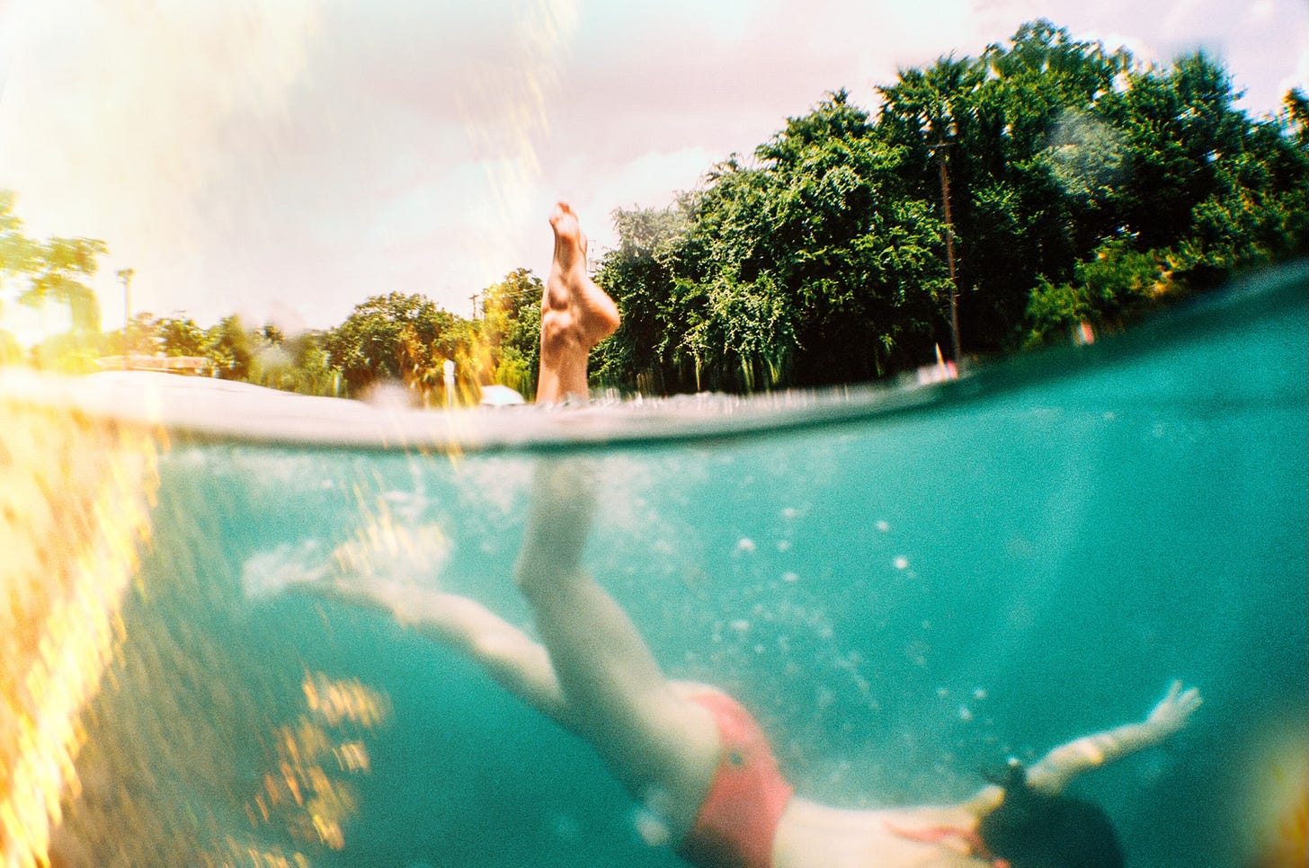 A split-level photo captures a swimmer in a red swimsuit diving underwater, with their legs breaking the surface. Sunlight and trees frame the vibrant outdoor scene.