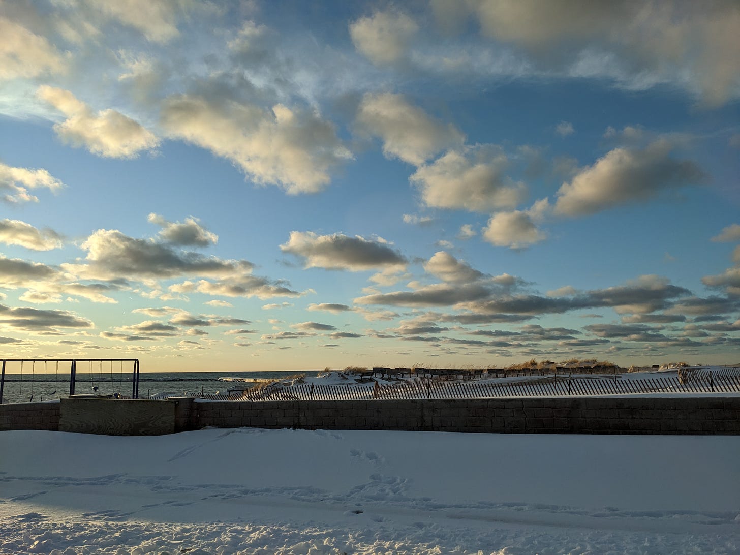 A photo of Lake Michigan in winter. There's white snow in the foreground and a low wall leading to the beach area. An empty swingset, snow fence, and a number of benches sit on the beach. The sky is many shades of blue with a number of clouds scattered throughout.