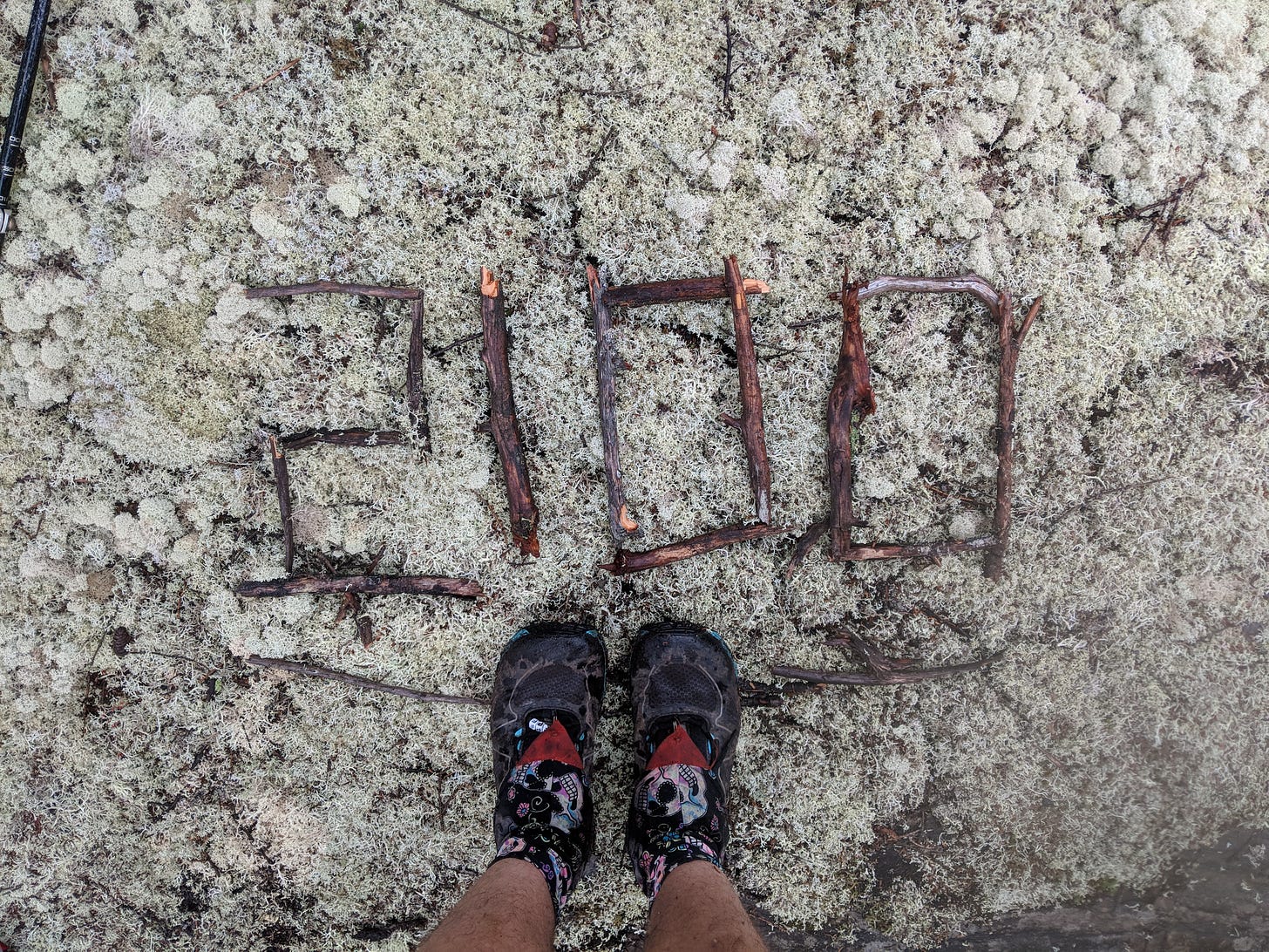 A photo of Sassafras' shoes. She's standing on a lichen-covered rock on the Appalachian Trail where someone has formed the number 2100 out of sticks.