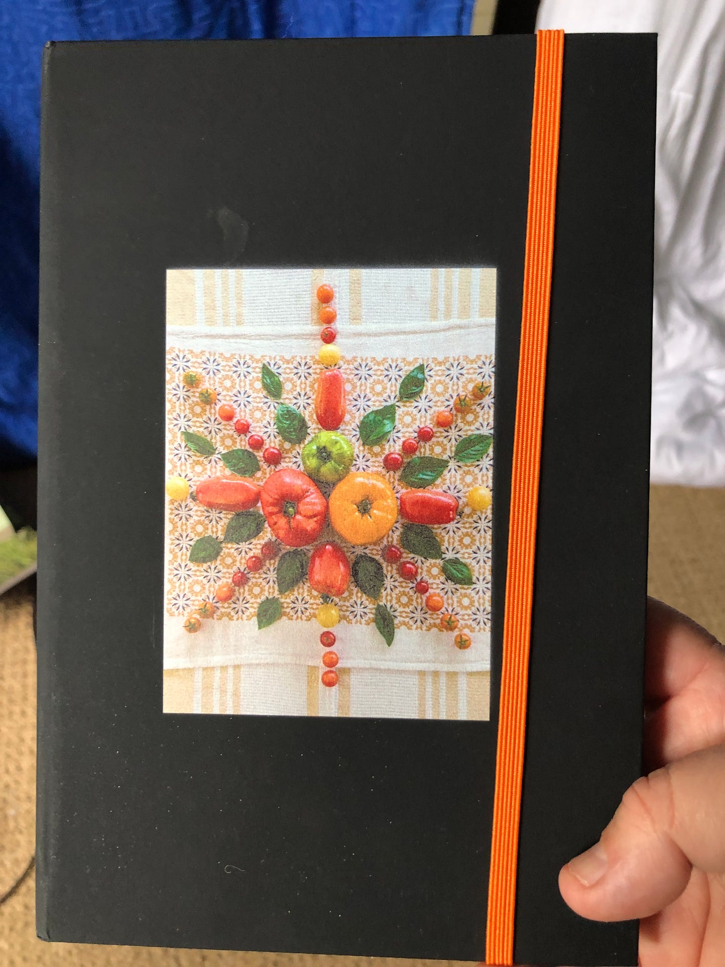 Black notebook with orange elastic band with an image of a harvest of tomatoes and basil leaves artfully arranged on a background napkin print