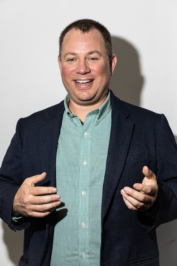 Mike Masnick smiles and gestures with both hands with his back to a white wall. He is wearing a blue blazer over a green checkered button-down shirt.