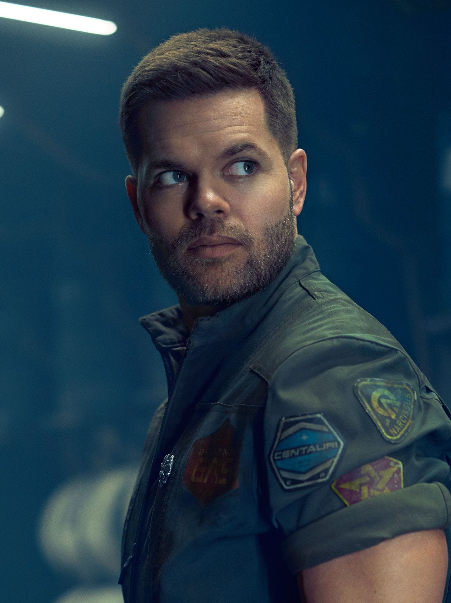 Still shot of Wes Chatham who plays Amos on The Expanse -- he's a white man with sandy brown hair, big eyes, stubble on his jaw, and a muscular form (sorry I don't know how else to phrase this). He's wearing one of the jumpsuits they always wear when they're crew on a ship.